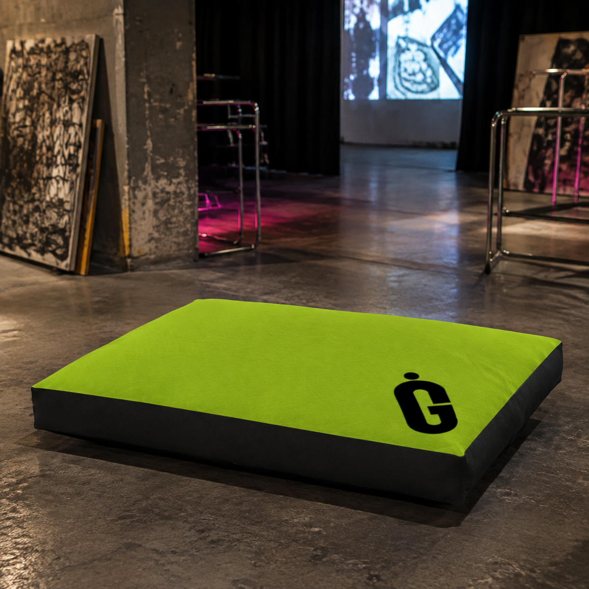 A rectangular floor cushion or pet bed with a bright lime green top and black sides, featuring a black stylized 'G' logo, placed on a concrete floor in an industrial gallery setting.
