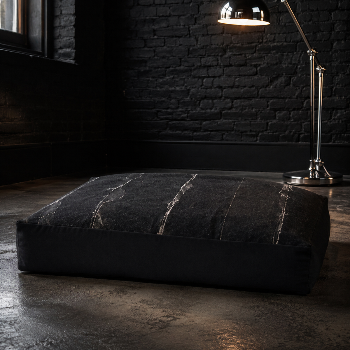 A large rectangular floor cushion in black fabric with a distressed, cracked texture, set on a dark concrete floor against a black brick wall with a lit industrial floor lamp.
