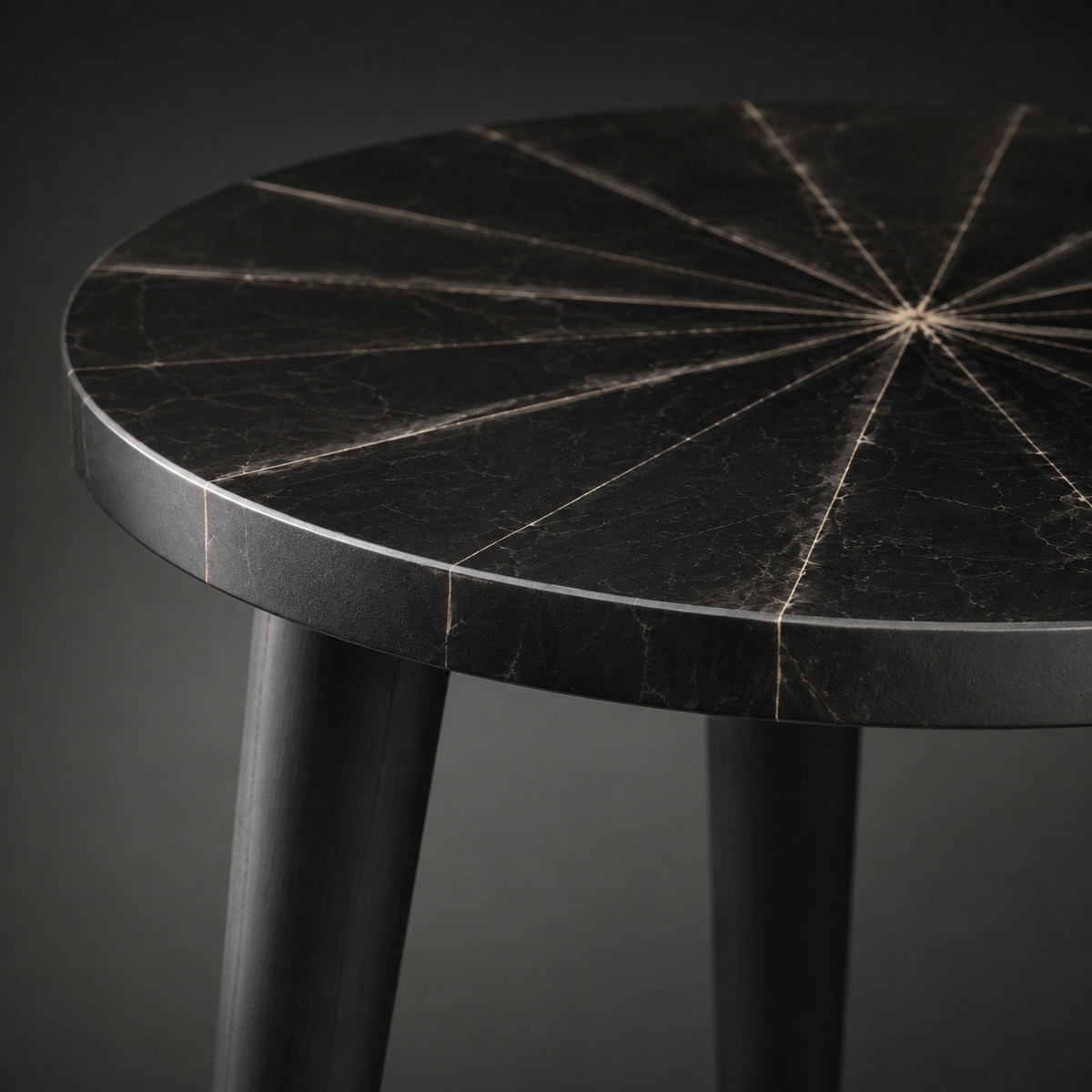 Close-up of a round black table with a segmented inlay pattern radiating from the center, supported by dark legs against a dark gray background.