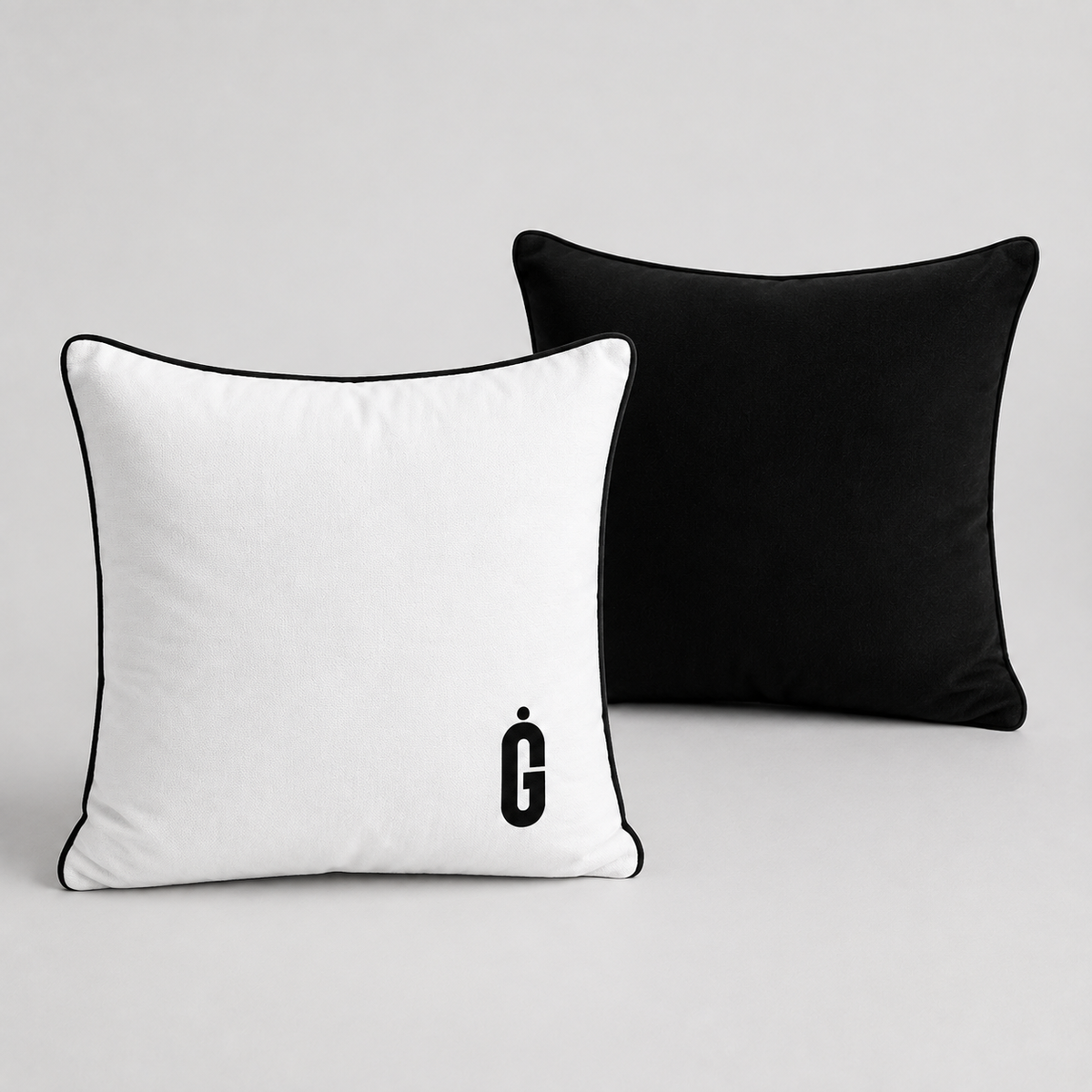 Two square throw pillows on a light gray background: one white pillow with black piping and a black monogram logo, and one black pillow behind it.