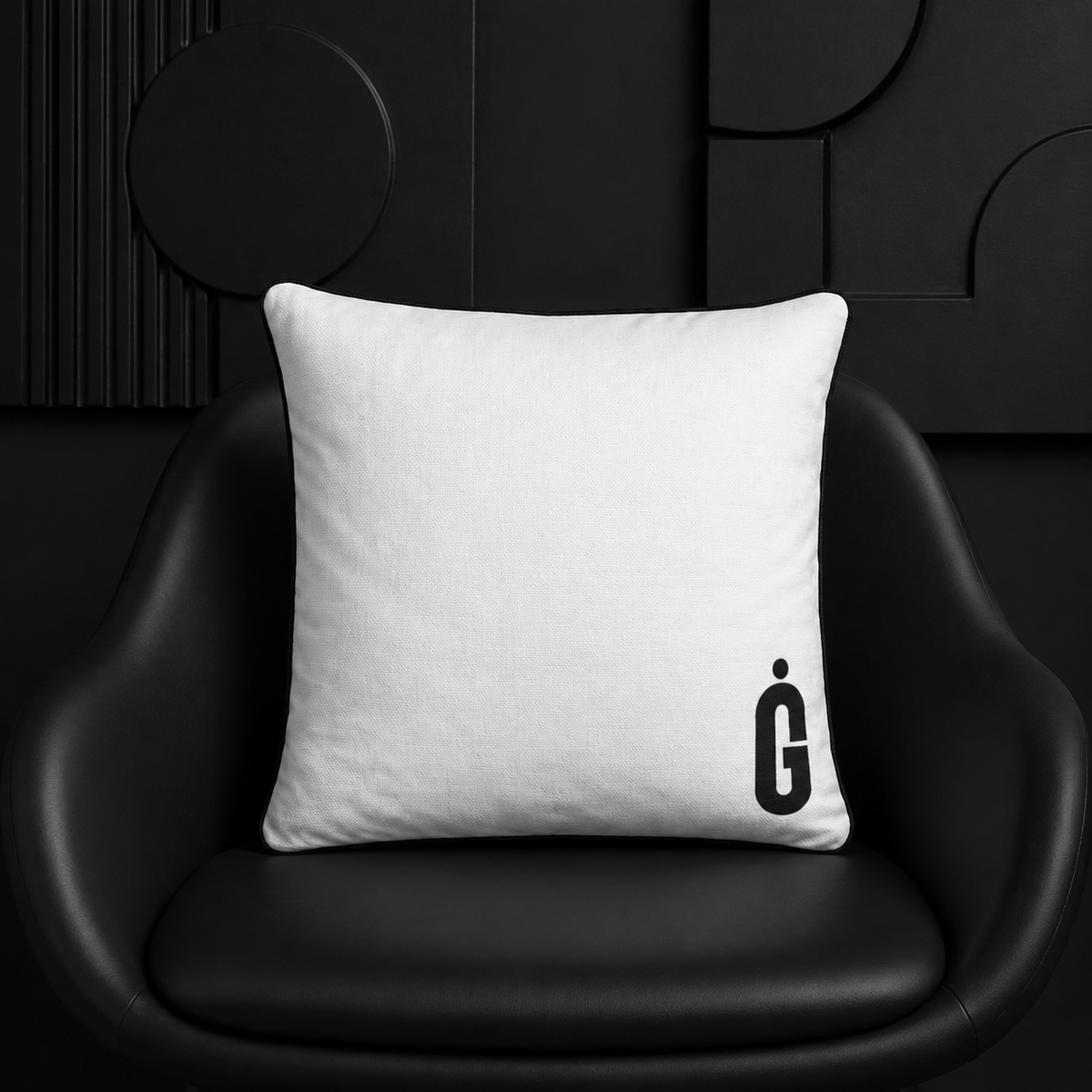 A square white throw pillow with a black stylized 'G' logo printed in the bottom right corner, resting on a black leather chair against a dark modern wall.