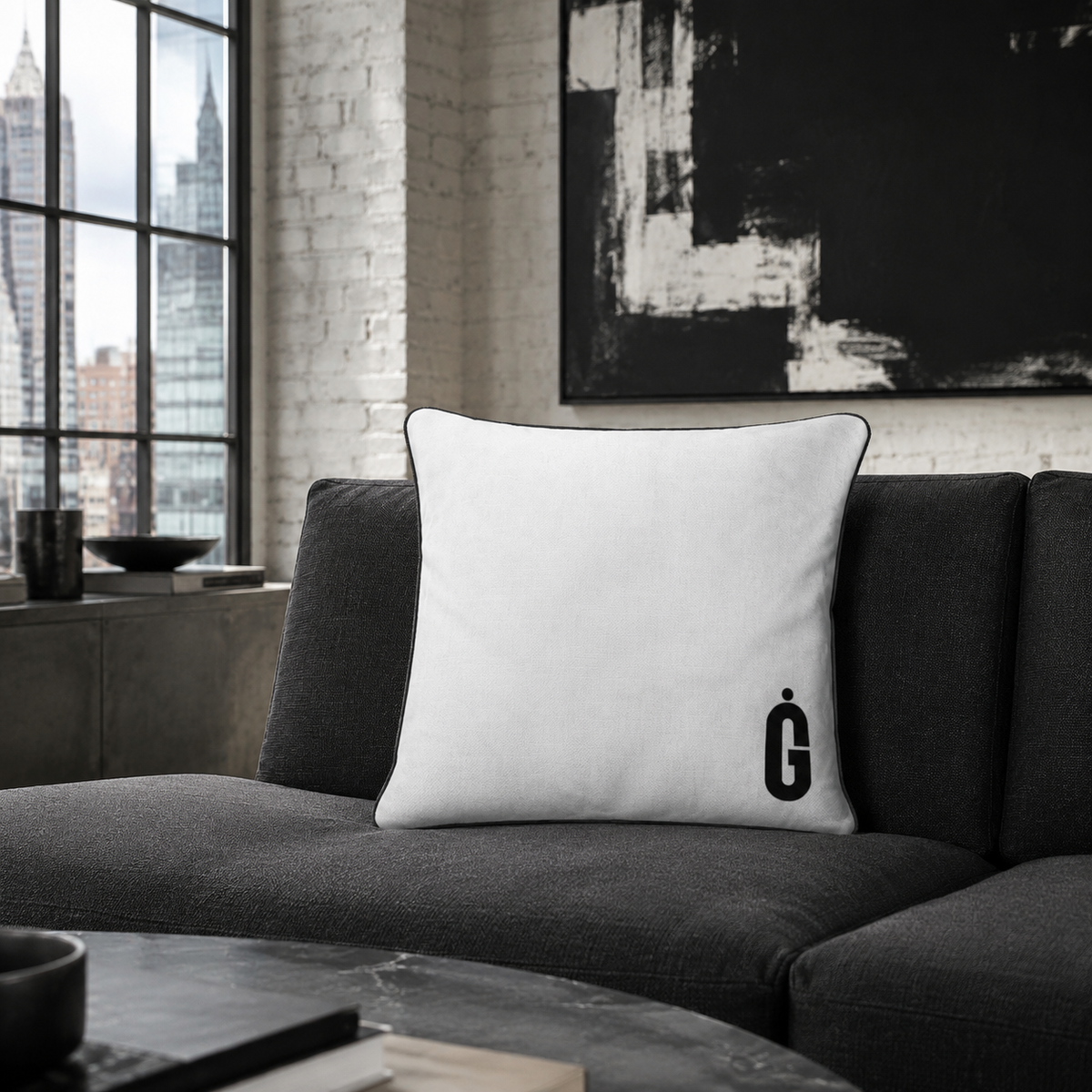 A square white throw pillow with a black border and a black monogram logo on the bottom right, resting on a dark gray sofa in a modern living room setting.